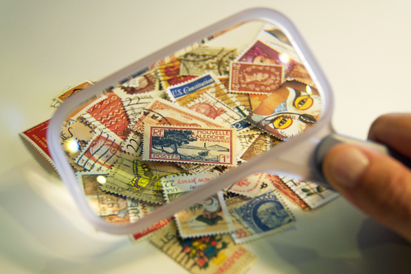 A hand holds a magnifying glass as a stamp collector examines a selection of postage stamps destined for an album.
